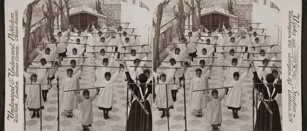 Sepia stereograph of rows of girls in white pinafores raising wooden wands in a courtyard, captioned Jaffa, Palestine, 1900