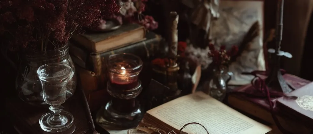 Candlelit writing desk with open old books, a pair of round spectacles, dried red flowers and a glass goblet.