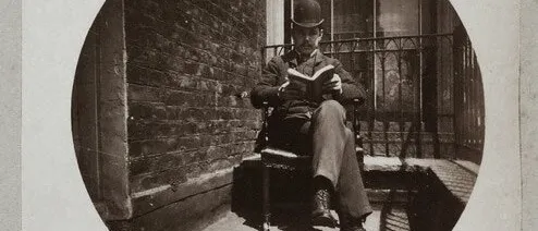 Sepia circular photograph of a man in a bowler hat reading a book in a chair on a balcony beside a brick wall