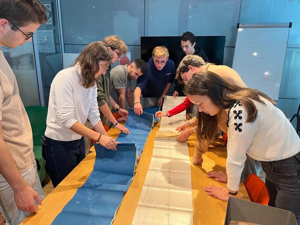 About nine young people lean over a long table, unrolling a strip of blue plans beside a long paper technical drawing ruled with red lines.