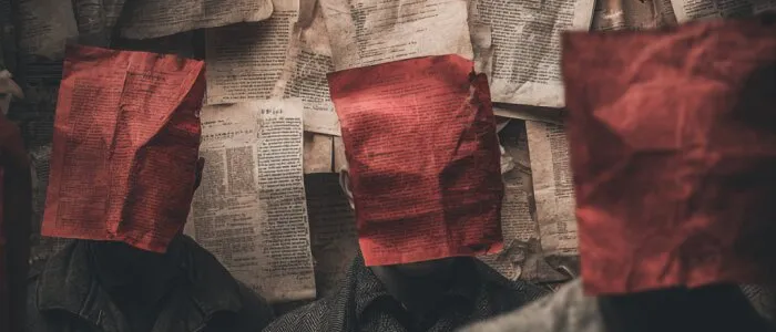 Three people in dark coats with crumpled red paper over their faces, before a wall of pinned newspaper pages