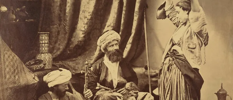 Sepia studio photograph: two turbaned men sit on rugs, one with a long-necked string instrument, watching a woman dance with raised arms.