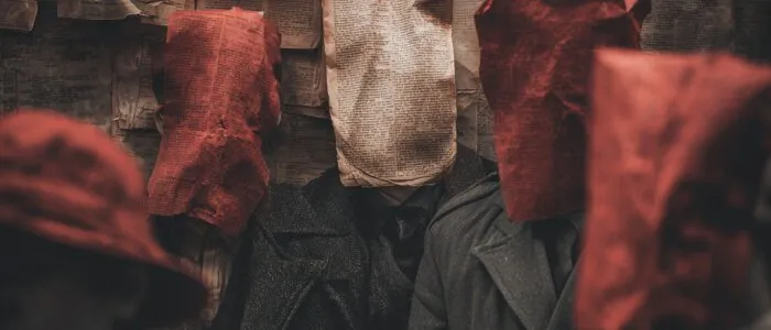 Figures in dark coats, their heads covered by red and cream newspaper bags, before a wall pasted with newsprint