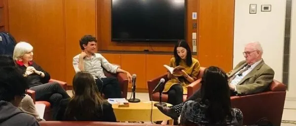 Four speakers in armchairs at a panel in a wood-panelled room, one reading from a book, with an audience in the foreground