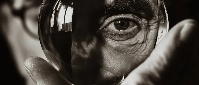 Black-and-white close-up of a hand holding a glass sphere that magnifies a man's eye, his glasses blurred behind