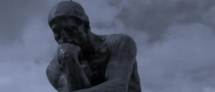 Dark bronze statue of a hunched, seated figure with its chin resting on one hand, against a grey-blue sky, with the red PICT mark in the lower centre