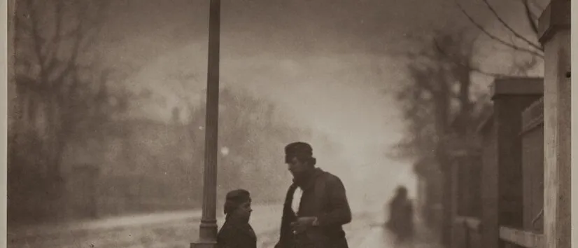 Sepia photograph of a boy and a man talking under a lit gas street lamp on a wet, misty pavement