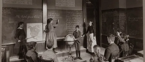 Old sepia photograph of a classroom: pupils at desks watch children at blackboards with a map and a globe
