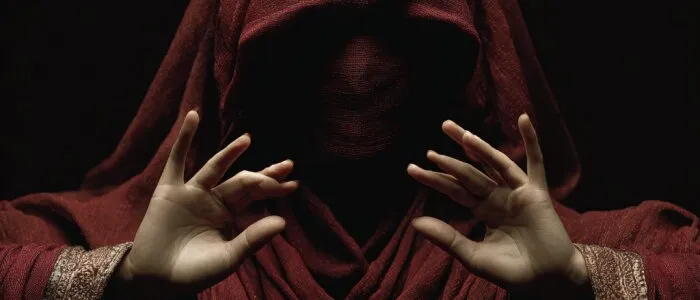 A figure draped in a dark red veil, face hidden in shadow, raising both hands against a black background