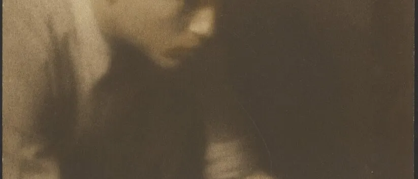 Soft-focus sepia photograph of a young man in a cap leaning over a table, writing a note